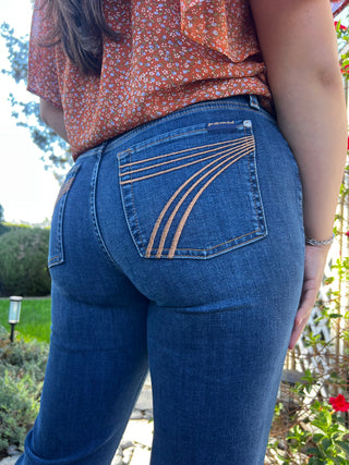 A woman wearing dark wash jeans with gold 7 stitching, showing the back pocket and the seam detail.