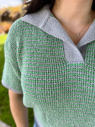 A woman wearing a short sleeve green and grey sweater with a collar while wearing blue jeans.