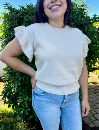 A woman wearing a cream short sleeve ruffle sweater paired with blue jeans, standing outdoors.