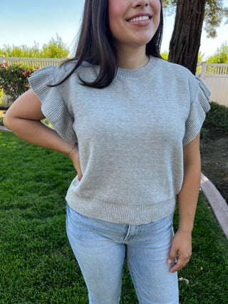 A woman wearing a grey short sleeve ruffle sweater paired with blue jeans, standing outdoors.