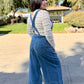 Woman wearing blue denim overalls with a white lace long sleeve and white sneakers standing 