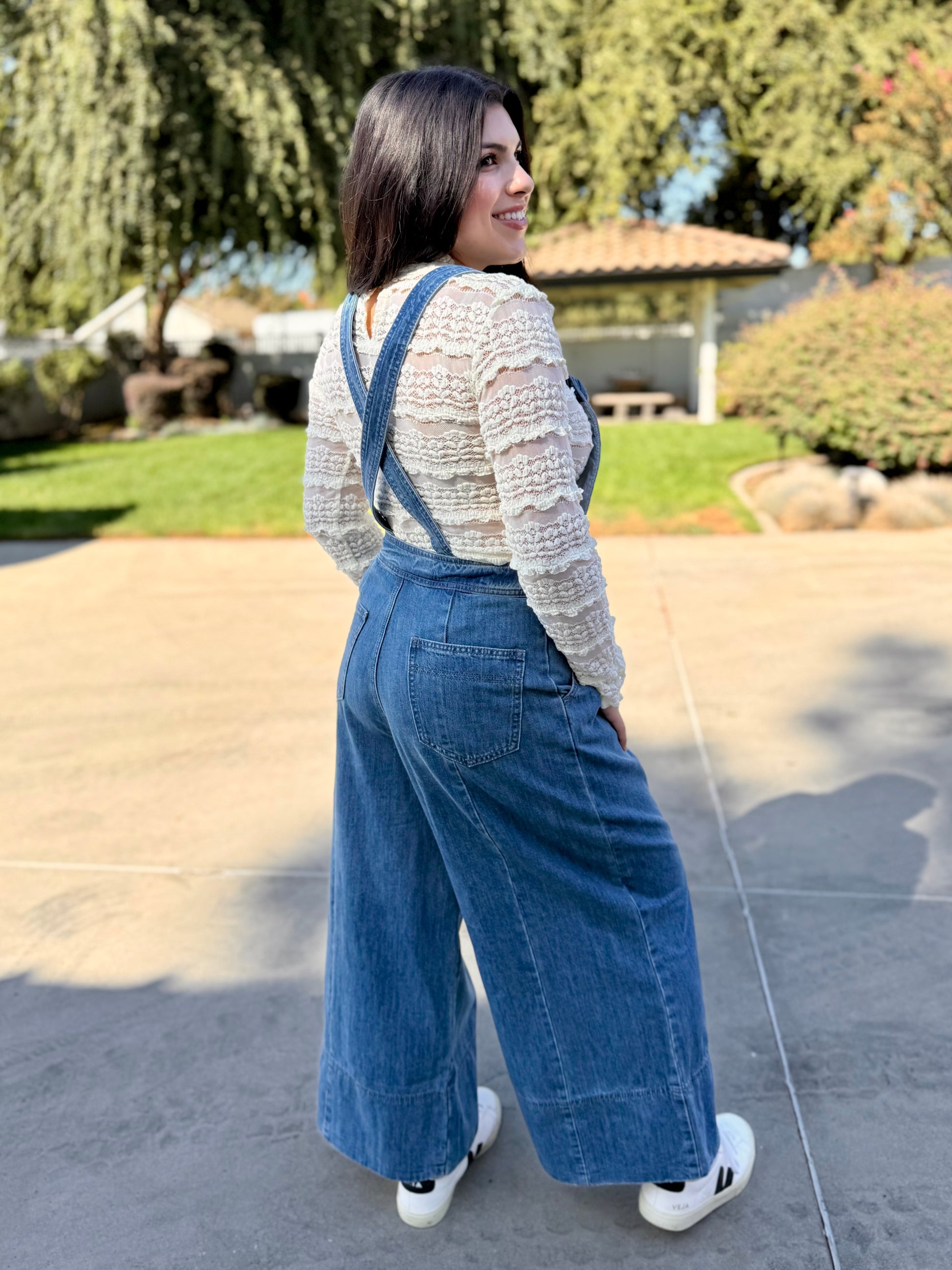 Woman wearing blue denim overalls with a white lace long sleeve and white sneakers standing 