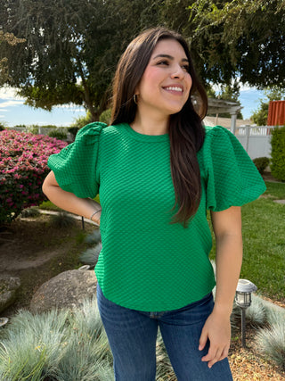 A woman wearing a green textured balloon sleeve top with a high neckline, paired with blue jeans.