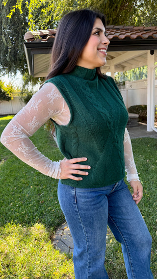 Woman wearing a green cable knit sleeveless sweater and blue jeans.
