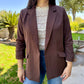 Woman wearing a brown blazer over a white lace top and blue jeans, standing outdoors.