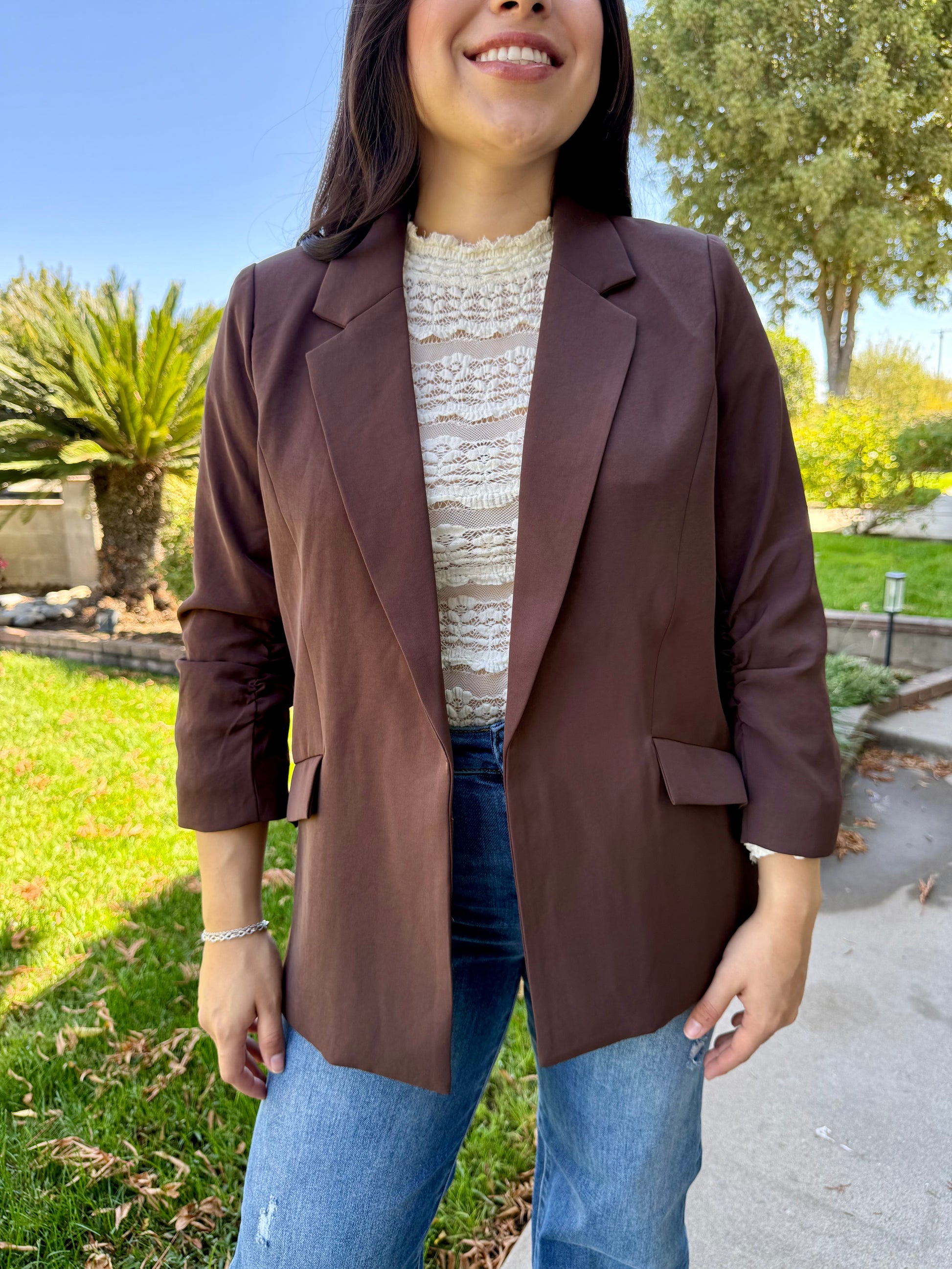 Woman wearing a brown blazer over a white lace top and blue jeans, standing outdoors.