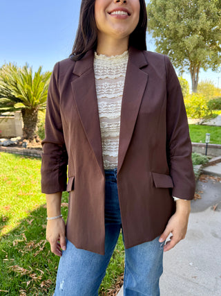 Woman wearing a brown blazer over a white lace top and blue jeans, standing outdoors.