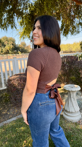 Woman wearing a brown top knitted top with a satin tie on the back and blue jeans.