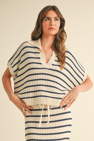 A woman wearing a striped collared blouse with short sleeves, wearing blue jeans. The blouse is beige and navy striped, and the photo is taken outdoors with flowers in the background.