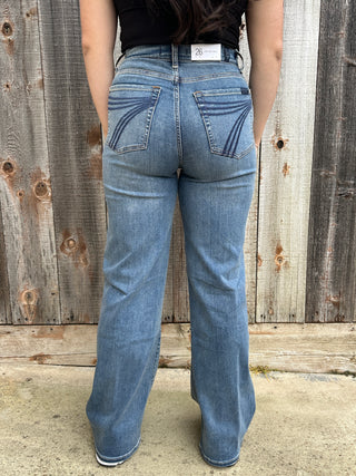 A woman wearing high waisted medium wash jeans in a wide leg style with a blue 7 stitching on the back pocket. 