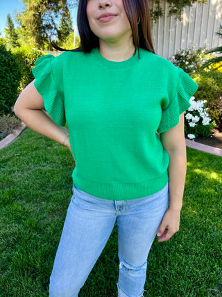 A woman wearing a green short sleeve ruffle sweater paired with blue jeans, standing outdoors.