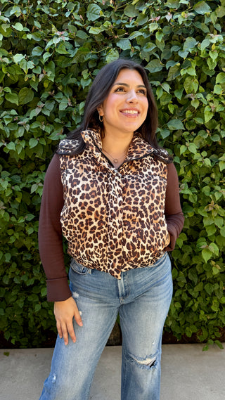 Woman wearing a leopard print vest over a brown long-sleeve shirt with blue jeans, standing against a green leafy background.