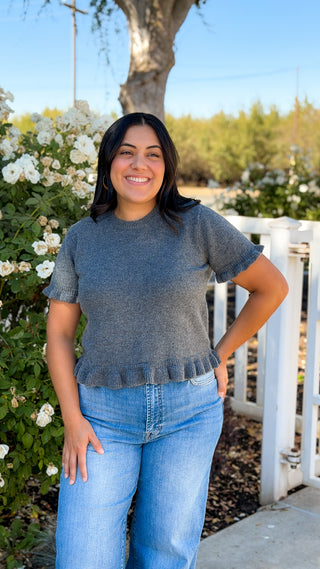 Model wearing charcoal gray short sleeve sweater top with ruffle hem and light wash jeans outdoors, front view