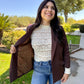 Woman wearing a brown blazer over a white lace top and blue jeans, standing outdoors.