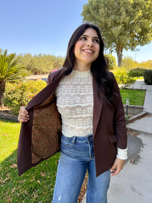 Woman wearing a brown blazer over a white lace top and blue jeans, standing outdoors.