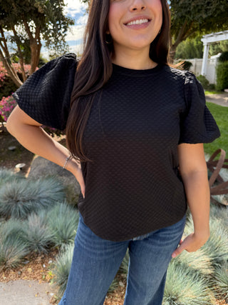 A woman wearing a black textured balloon sleeve top with a high neckline, paired with blue jeans.