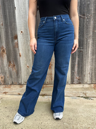 A woman wearing dark wash high waisted, wide leg jeans with a white 7 stitched on the back pocket. 