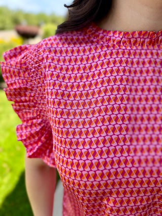 A woman wearing a red and pink patterned retro-inspired blouse with a ruffled high neckline and ruffle statement sleeves, paired with blue jeans and black shoes.