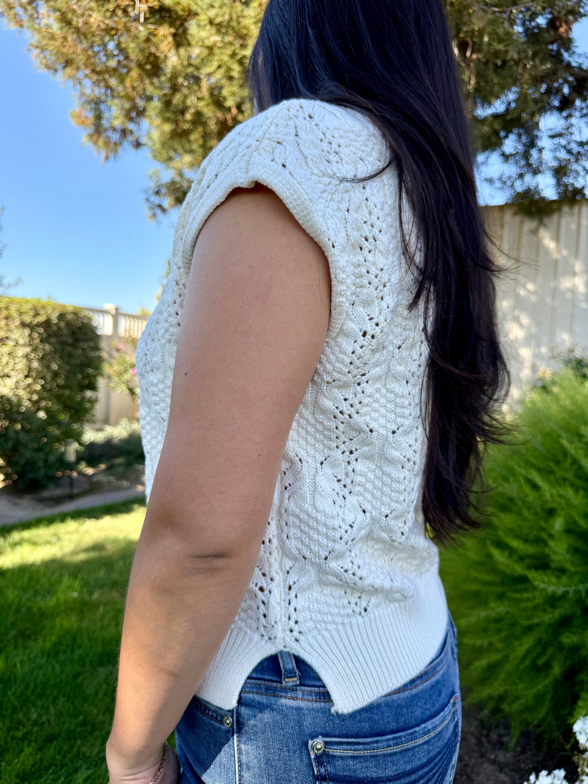 Person wearing a white sleeveless knitted top outdoors with greenery in the background