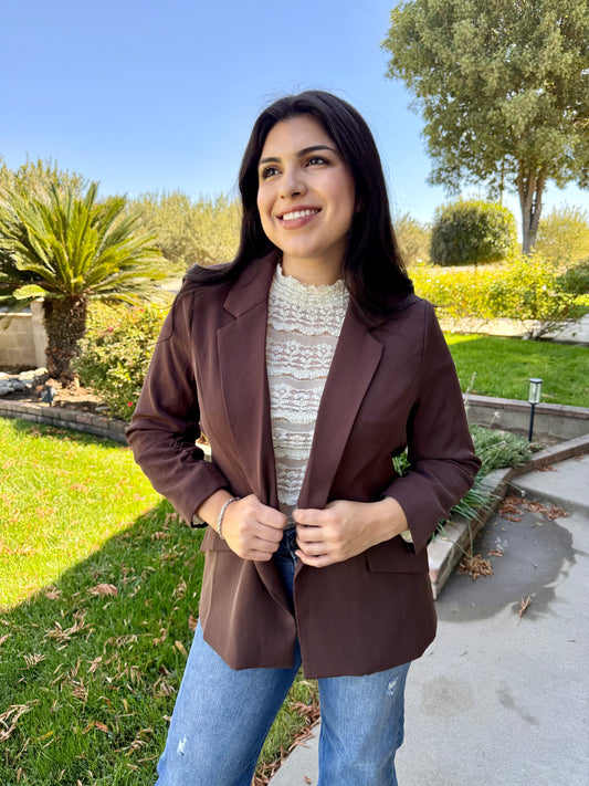 Woman wearing a brown blazer outdoors with greenery in the background