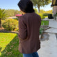 Person wearing a brown coat standing on a sidewalk with trees and grass in the background