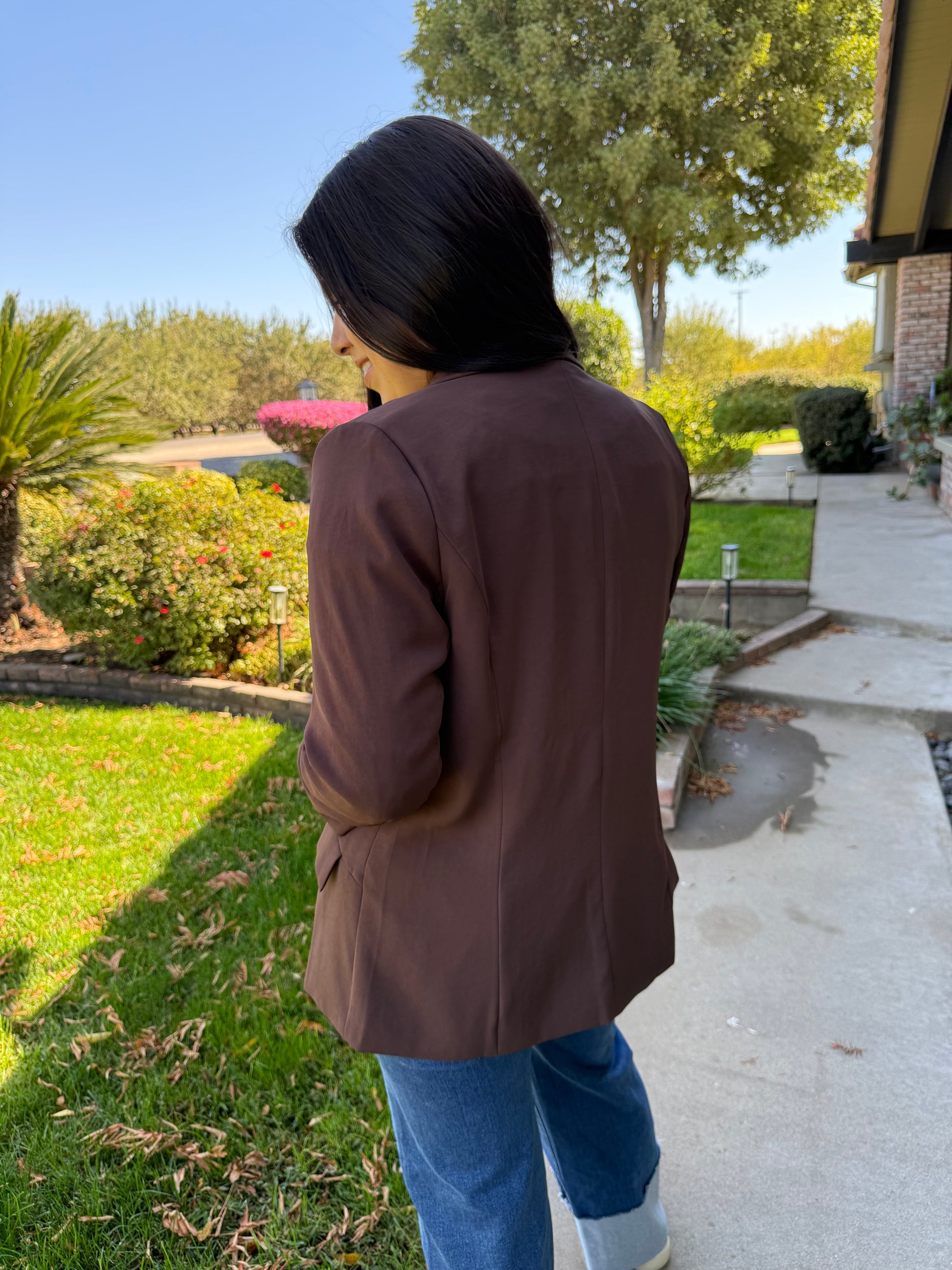 Person wearing a brown coat standing on a sidewalk with trees and grass in the background