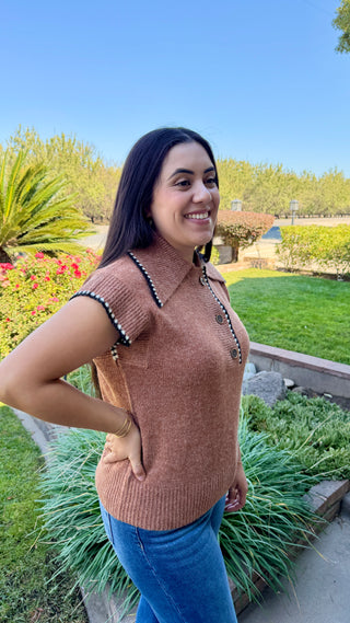 Woman wearing a brown sleeveless sweater with decorative black and white trim.