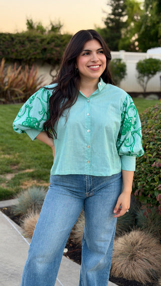 Woman wearing a green blouse with green patterns and jeans standing outdoors.
