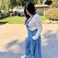 Woman wearing blue denim overalls with a white lace long sleeve and white sneakers standing 