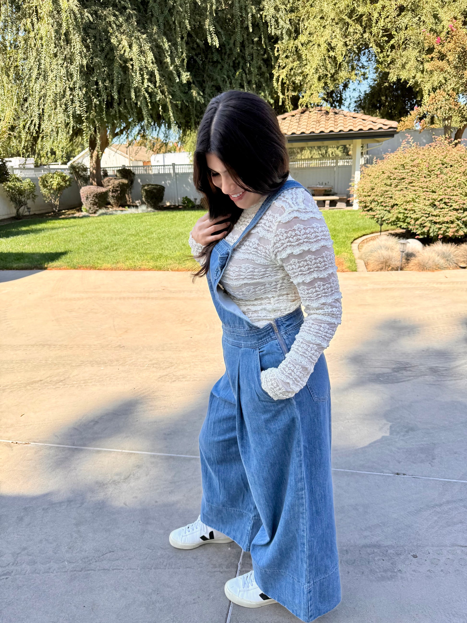 Woman wearing blue denim overalls with a white lace long sleeve and white sneakers standing 