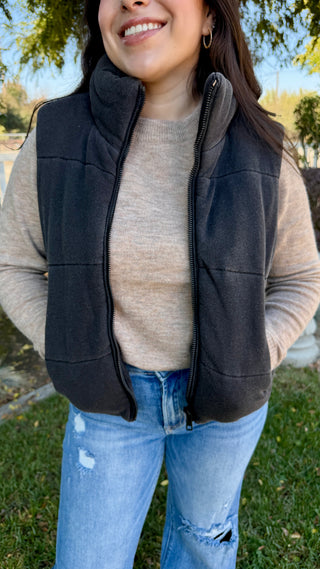 Woman wearing a black vest over a beige sweater with blue jeans outdoors.