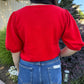 Person wearing a red top and blue jeans standing outdoors with greenery in the background