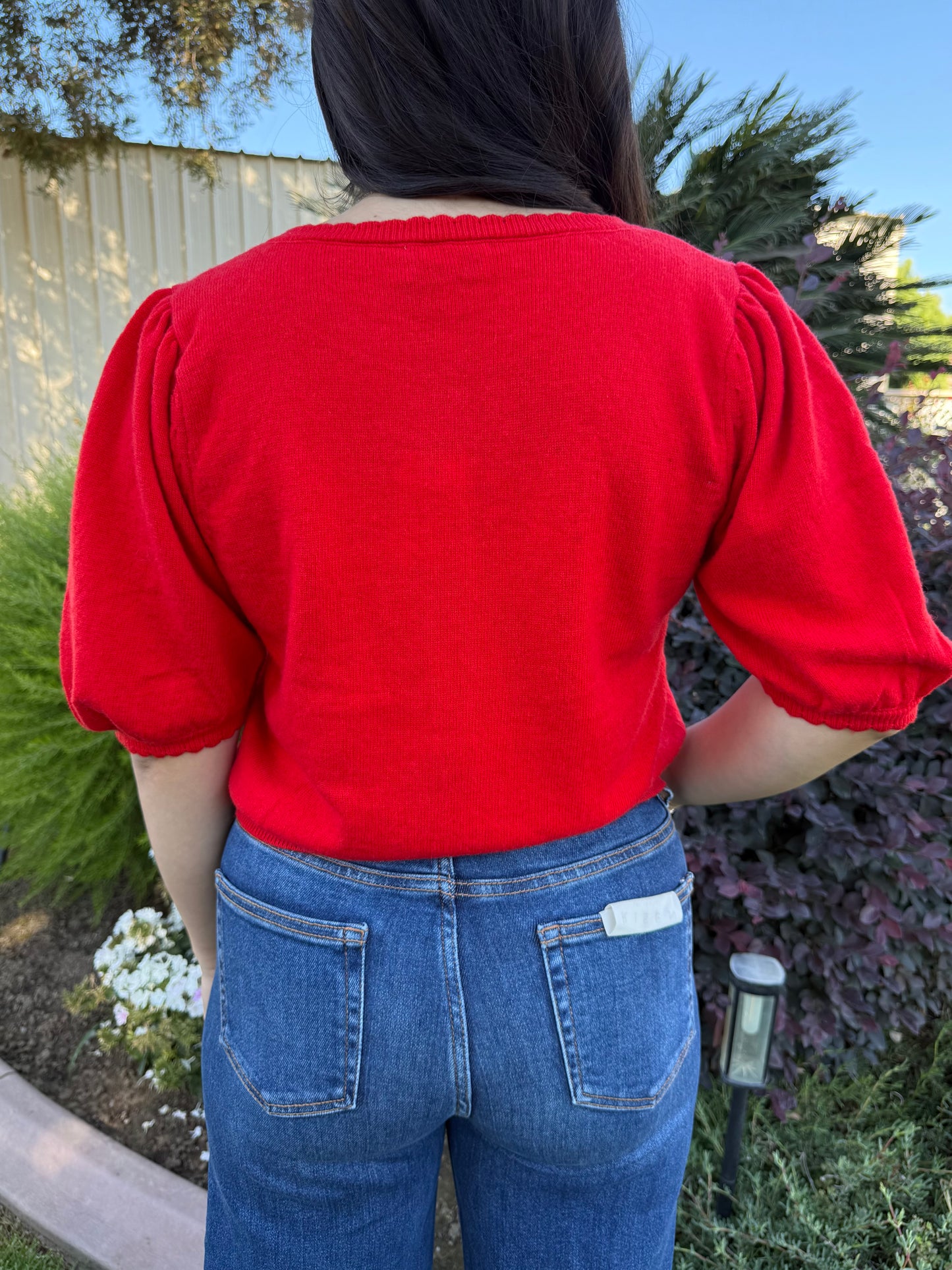 Person wearing a red top and blue jeans standing outdoors with greenery in the background