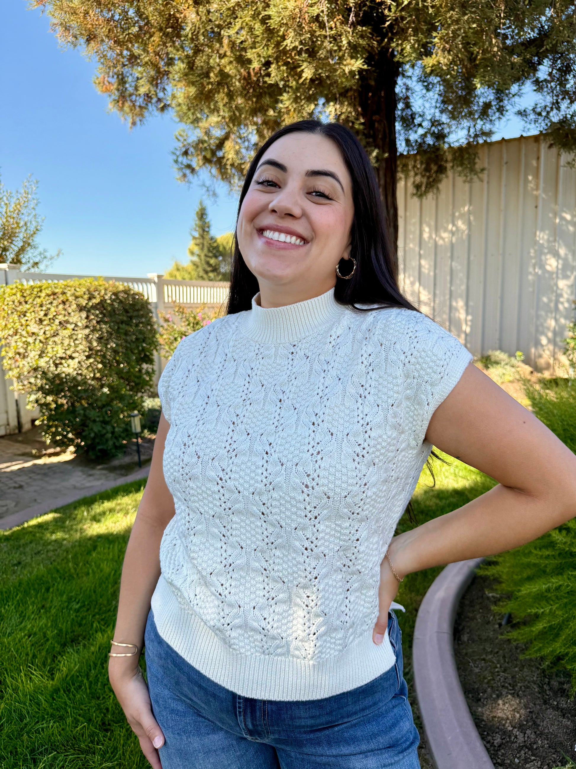 Woman wearing a light blue knitted top outdoors with trees and a fence in the background