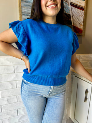 A woman wearing a blue short sleeve ruffle sweater paired with blue jeans, standing outdoors.