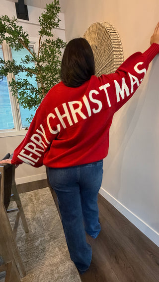 Person wearing a red sweater with 'Merry Christmas' text, standing in a room with a plant and window.