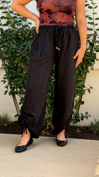 Front view of black wide leg drawstring pants with gold-tipped drawstring waist and ruched ankle ties cinched, worn with a burgundy print top and black flats