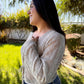Woman wearing a beige cardigan and blue jeans standing outdoors with greenery in the background