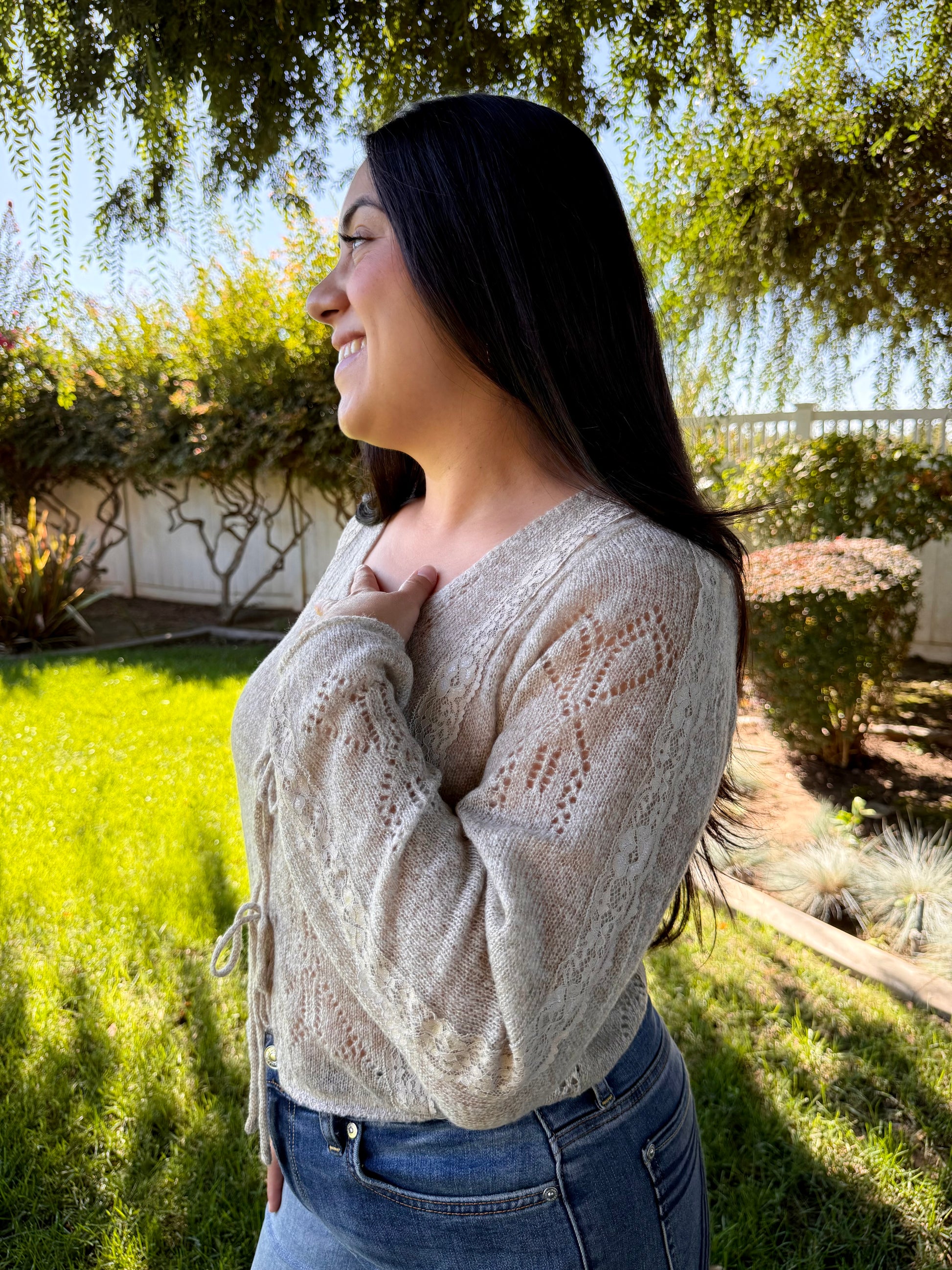 Woman wearing a beige cardigan and blue jeans standing outdoors with greenery in the background