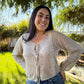 Woman wearing a beige cardigan and blue jeans standing outdoors with greenery in the background