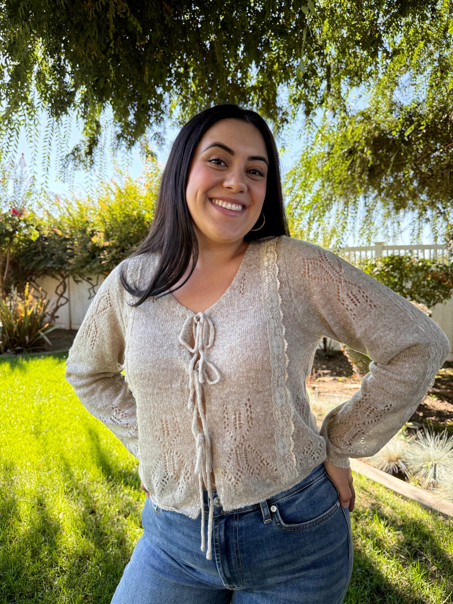 Woman wearing a beige cardigan and blue jeans standing outdoors with greenery in the background