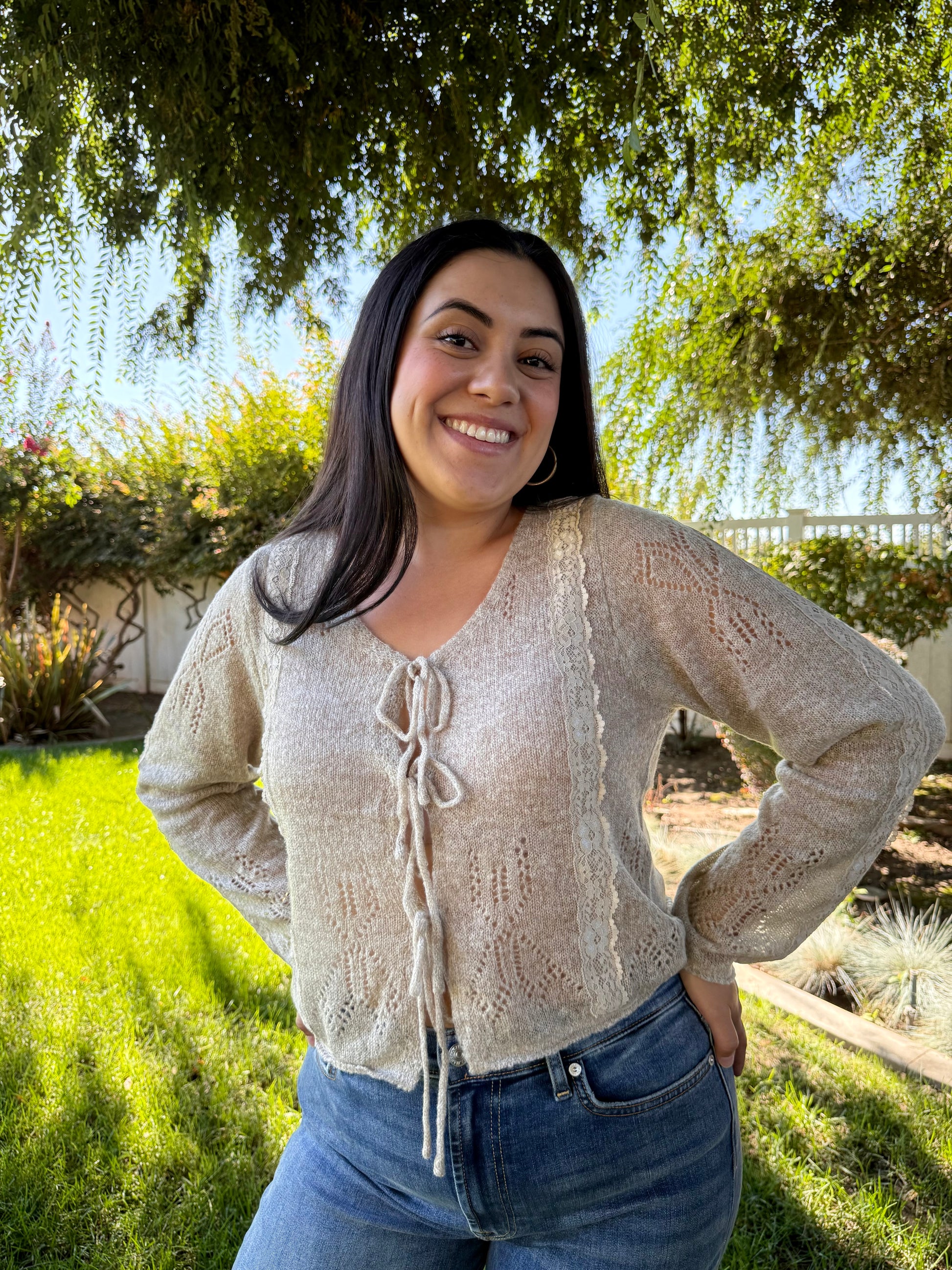 Woman wearing a beige cardigan and blue jeans standing outdoors with greenery in the background
