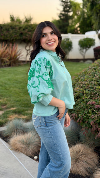 Woman wearing a light green blouse with embroidery and blue jeans standing outdoors.