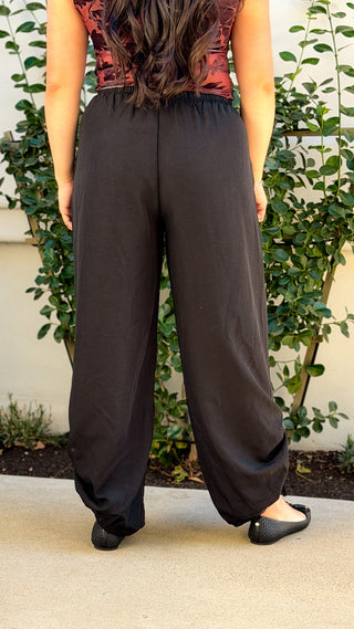 Back view of black wide leg drawstring pants with elastic waistband and ruched ankle ties, worn with black flats