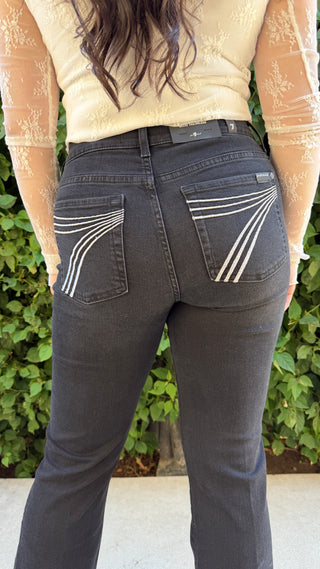Person wearing black jeans with white stitching, standing against a green leafy background