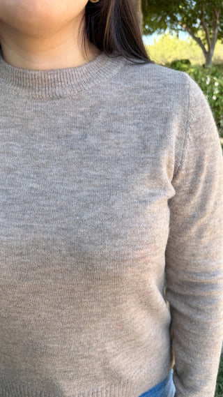 A woman wearing a long sleeve beige sweater with blue jeans outdoors.