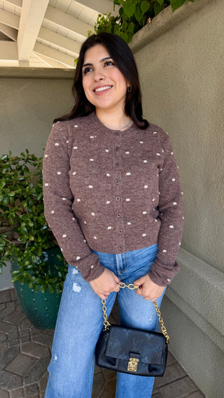 Woman wearing a brown polka dot sweater and blue jeans, holding a black handbag.
