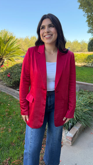 Woman wearing a long sleeve red satin blazer and blue jeans.