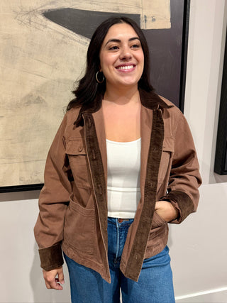 Woman wearing a brown jacket and blue jeans standing indoors with a neutral background